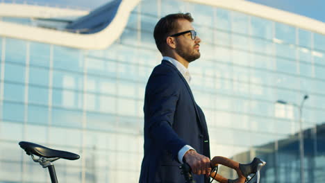 Portrait-shot-of-an-attractive-man-walking-with-his-bike-beside-next-to-a-big-building