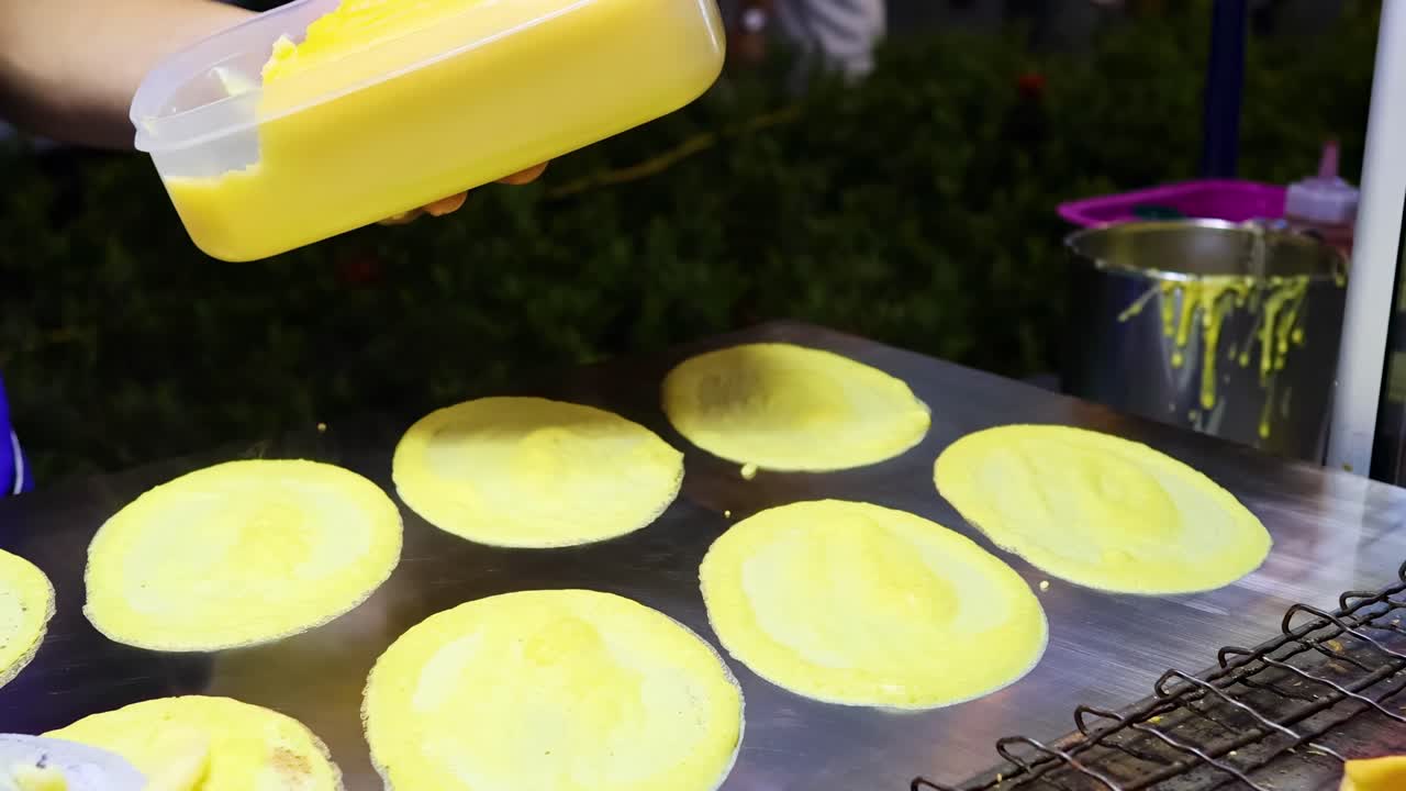A vendor skillfully prepares crispy pancakes on a street stall in Bangkok, using a batter dispenser under bright lighting