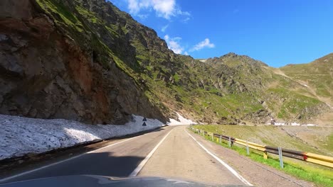 pov conductor en la carretera de montaña transfagarasan, rodeado de altos y verdes picos de montaña con manchas de nieve y un cielo azul claro