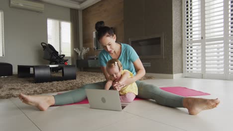 Caucasian-mother-holding-her-baby-practicing-yoga-on-yoga-mat-in-front-of-laptop-at-home