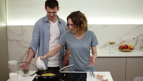 cheerful couplemaking breakfast together. girl pouring mixed eggs from a bowl to a hot pot on a kitchen stove. husband standing behind and watching. slow motion