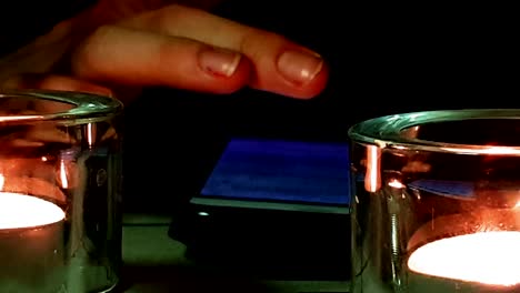 toma cercana, velas encendidas enfocadas y teléfono inteligente navegando con la mano en segundo plano, mujer siguiendo tristes actualizaciones de noticias