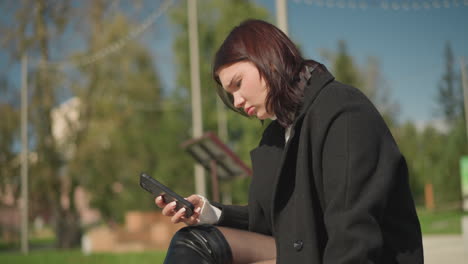 woman seated outdoors looking focused on phone in her hand, ring on hand, with a blurred background of greenery and trees in a peaceful, sunny environment