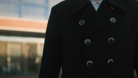 man in black suit with shining metallic buttons stands outdoors with a composed expression as sunlight reflects off his right hand, exuding professionalism, background features modern glass building