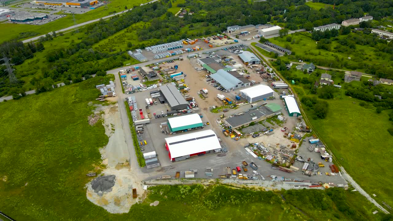 High altitude aerial drone footage orbiting around a countryside car park or an idustrial area that is fenced off from a highway and summwe green nature around it. Big warehouses and rooftops visible