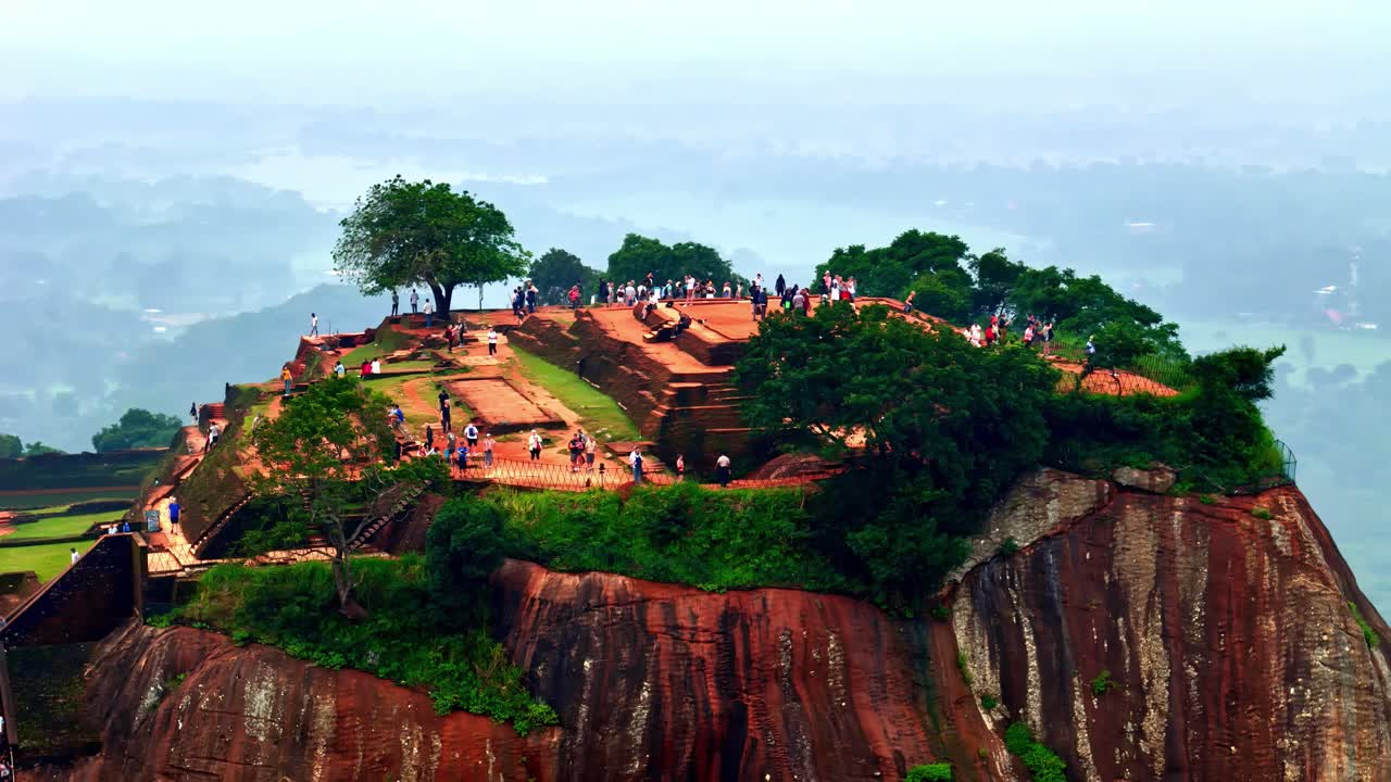 Drone aerial view of Sri Lanka's Sigiriya rock fortress with ancient frescoes, capturing the top of the plateau with ruins, gardens, and tourists under clear sky, highlighting the UNESCO heritage site
