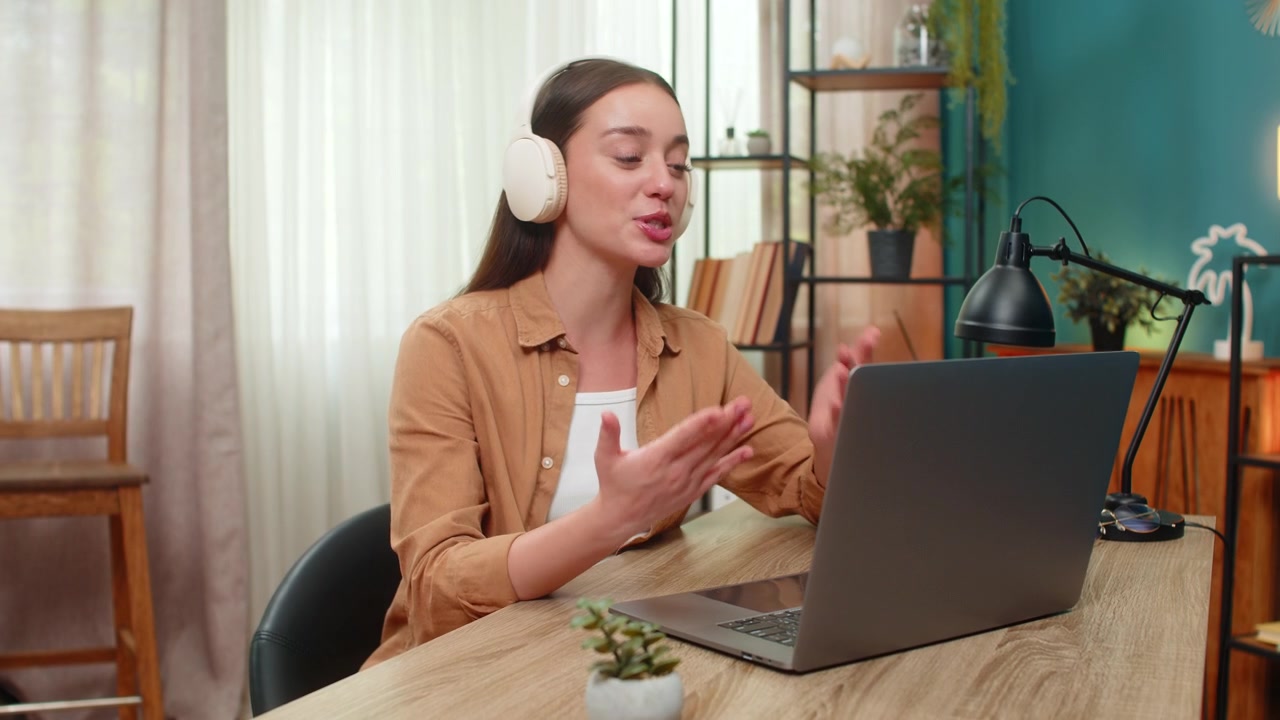 Young woman on laptop video call wearing wireless headset speaking live providing customer support