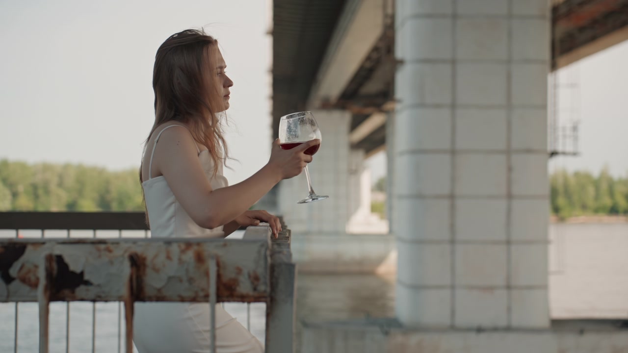 Donna in abito bianco sta in piedi su una vecchia struttura metallica arrugginita sotto un ponte, mentre tiene in mano e beve da un bicchiere di vino rosso. Lo sfondo presenta acqua e vegetazione con illuminazione diurna soffusa.