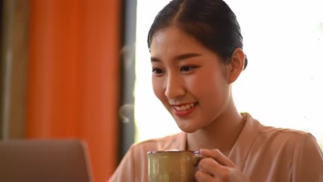 a young woman smiling while enjoying a warm drink and engaging with her laptop in a bright, cozy environment full of natural light