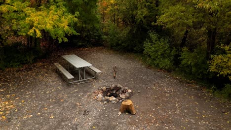 picnic table view backing up to reveal a rock ring fire pit at an empty campsite as the drone flys up showing how beautiful the autumn fall orange yellow trees are in october november