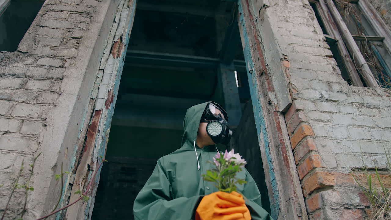 Man with gas mask holding flowers. Person in safety suit with flowers sitting in the abandoned building. Ecology pollution concept. Consequences of chemical attack.