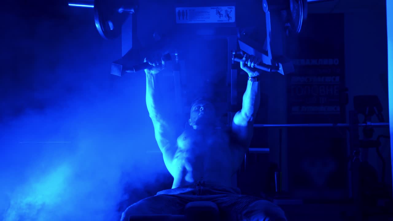 Sportsman training on simulator. Shirtless athlete doing his workout in the sports club. Handsome bodybuilder in the gym. Blue light filter and smoke.