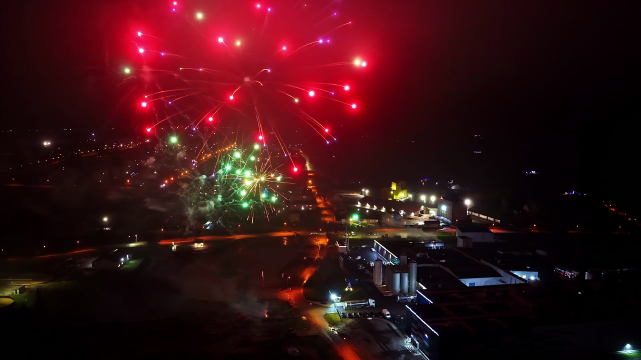 Drone aerial view of fireworks exploding above a city at night, with red smoke and glowing streetlights below. Festive holiday atmosphere over urban buildings.