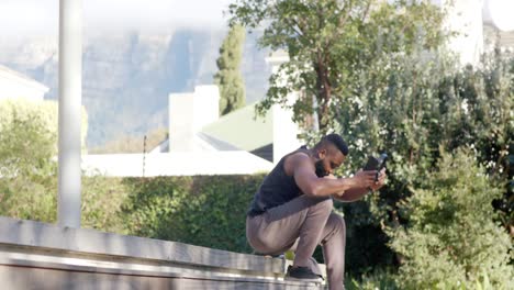 african american man resting using smartphone and headphones on deck in sunny garden, slow motion