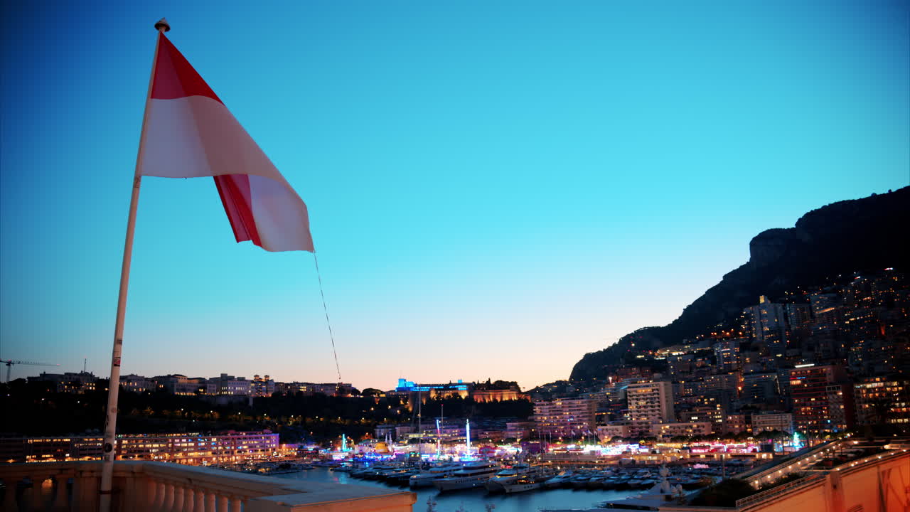 The flag of Monaco waving with the Port Hercule Funfair in Monaco in the evening on the background