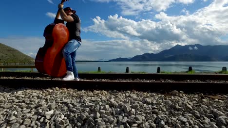 man playing double bass on train tracks by a lake