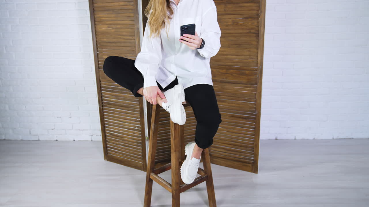 Young long-haired woman sits on wooden stool showing white shoes. Lady in shirt and trousers demonstrates a pair of shoes and uses phone.