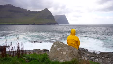 Weitwinkelaufnahme-Eines-Mannes-Mit-Gelbem-Regenmantel,-Der-Auf-Einem-Felsen-Sitzt-Und-Das-Färöische-Meer-Bewundert