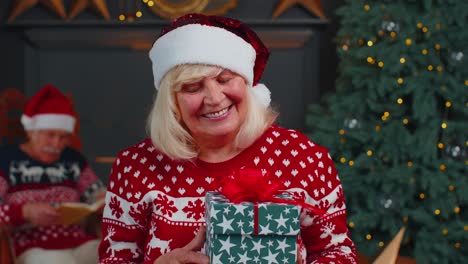 Elderly-grandmother-in-festive-sweater-presenting-Christmas-gift-box,-smiling,-looking-at-camera