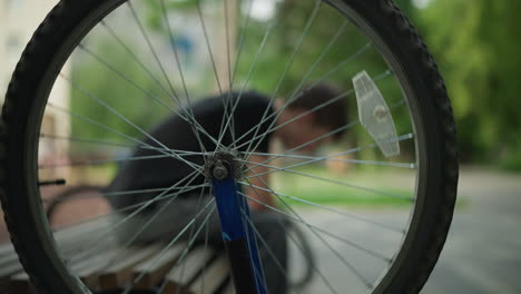 close-up of rotating bicycle wheel in focus, creating blur effect of a person seated on bench with the head bowed low, the background, including the figure and scenery, is blurred