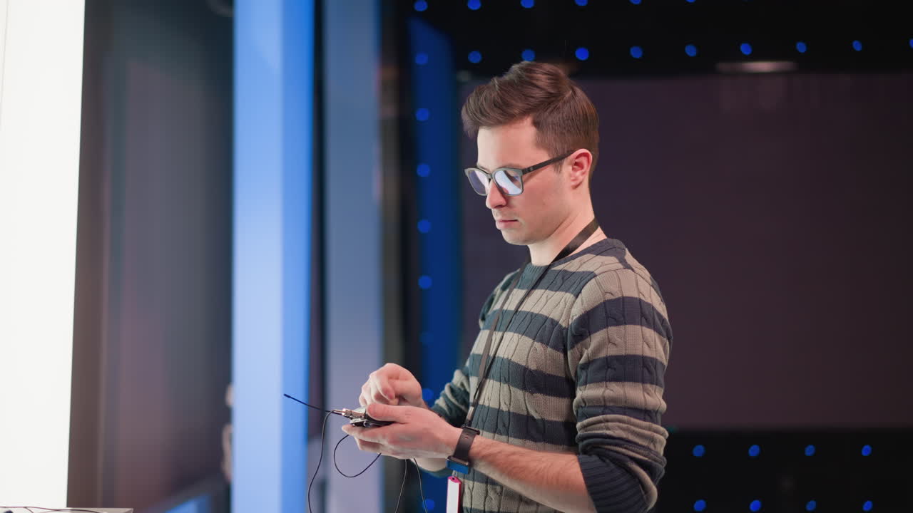 male technician wearing glasses adjusts wireless microphone transmitter cables while monitoring audio device inside television studio lit by blue backdrop lights during live broadcast prep