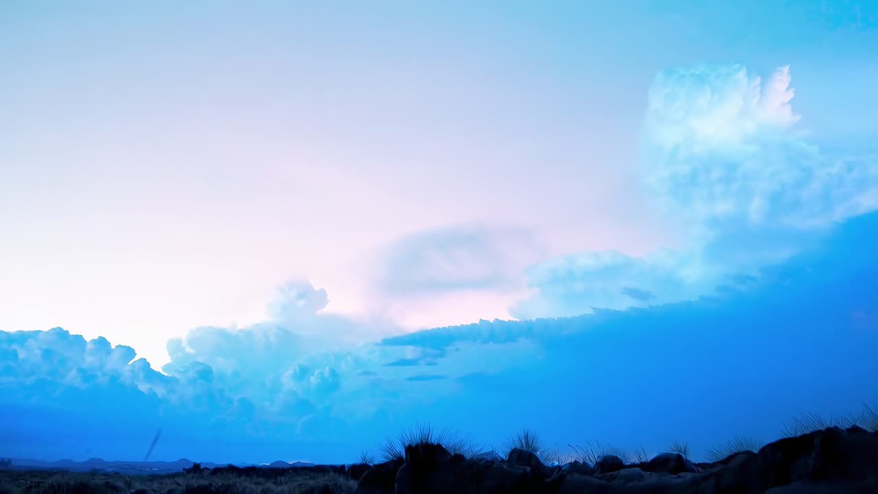 Una ripresa video grandangolare di un cielo notturno stellato con nuvole drammatiche e un orizzonte luminoso.