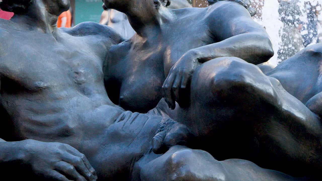 Bronze male and female statues appear to converse beside splashing fountain, modern urban background visible