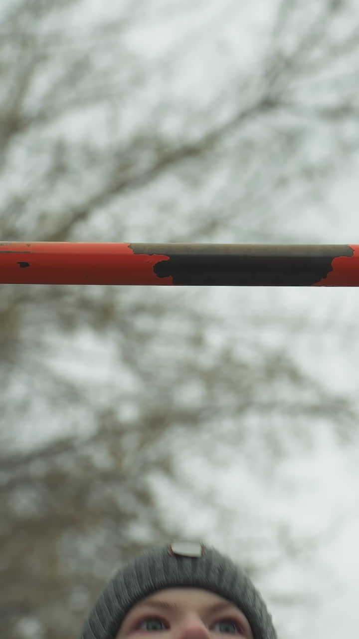 Close-up of a boy working out on pull-up bars in an outdoor gym area, after successfully completing his third pull-up, he struggles to continue, with a bare tree in the background