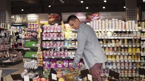 a smiling caucasian man in blue shirt shopping for fruits and vegetables in produce department of a grocery store supermarket. taking pomelo fruit, putting it into shopping trolley and walk further. big grocery shelves on the background