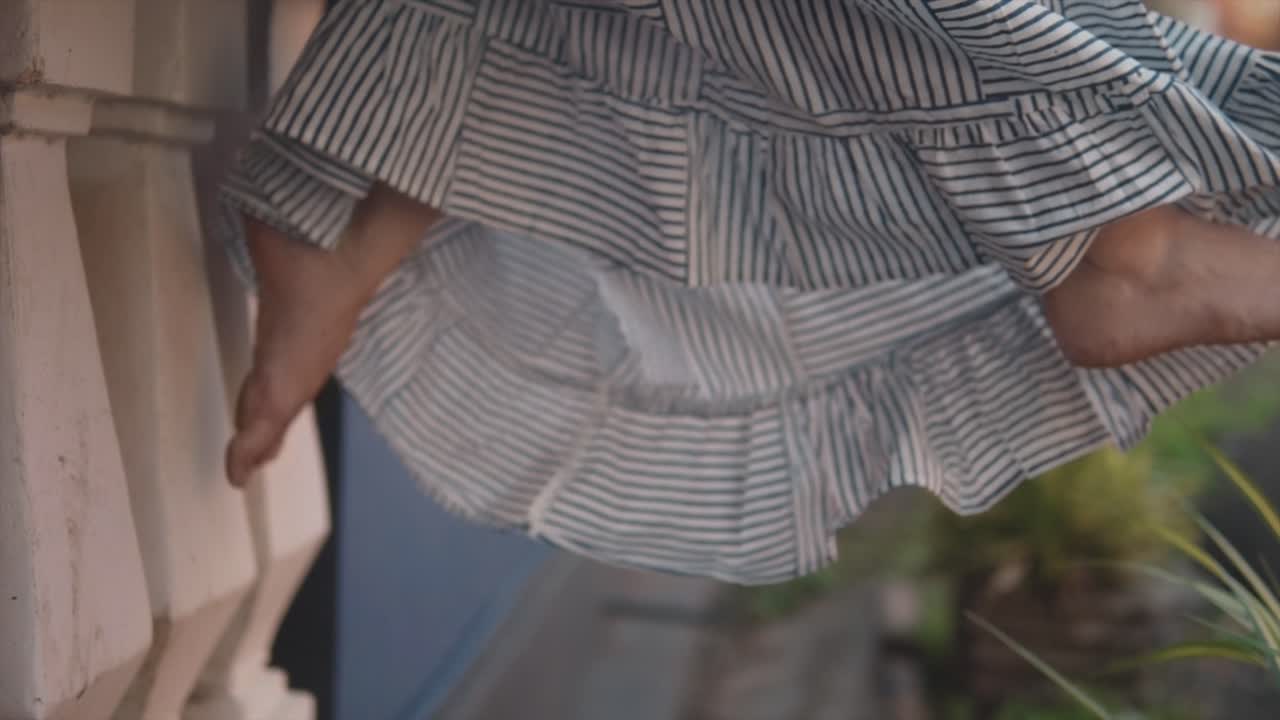 긴 여름 드레스를 입고 발코니에 앉아 자유롭게 흔들리는 젊은 여성의 발을 닫습니다