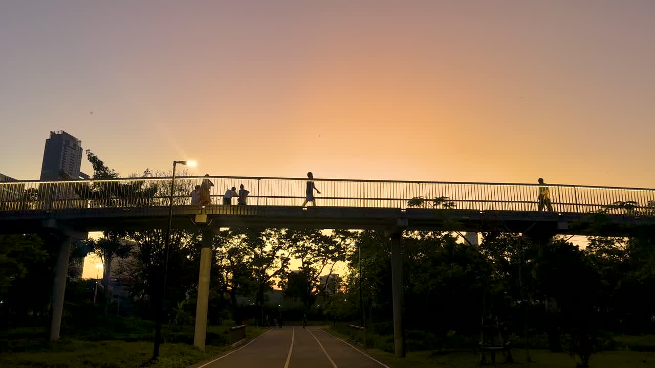 Silhouetted figures walk across a bridge at sunset in Benjakitti Park, Bangkok, with warm lighting and a tranquil atmosphere