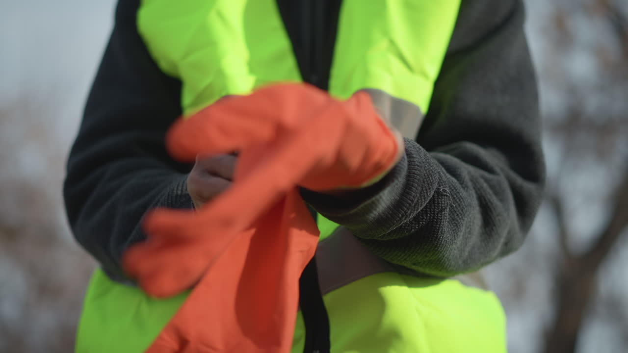 Worker in high visibility vest and dark sweater putting on bright orange protective gloves outdoors, ensuring safety and readiness for industrial or construction work in outdoor environment