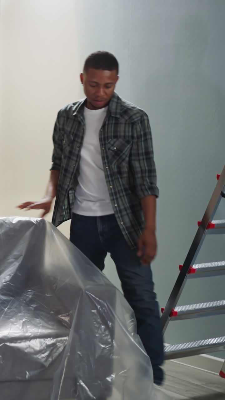 Black men cover new sofa with foil to protect from paint drops and sit down in room. Brothers prepare room for repairing. Home reconstruction organization