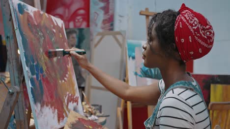 Close-Up-Of-Young-Woman-Painter-In-Painting-On-Easel-With-Paints-And-Brush-In-Cozy-Messy-Studio