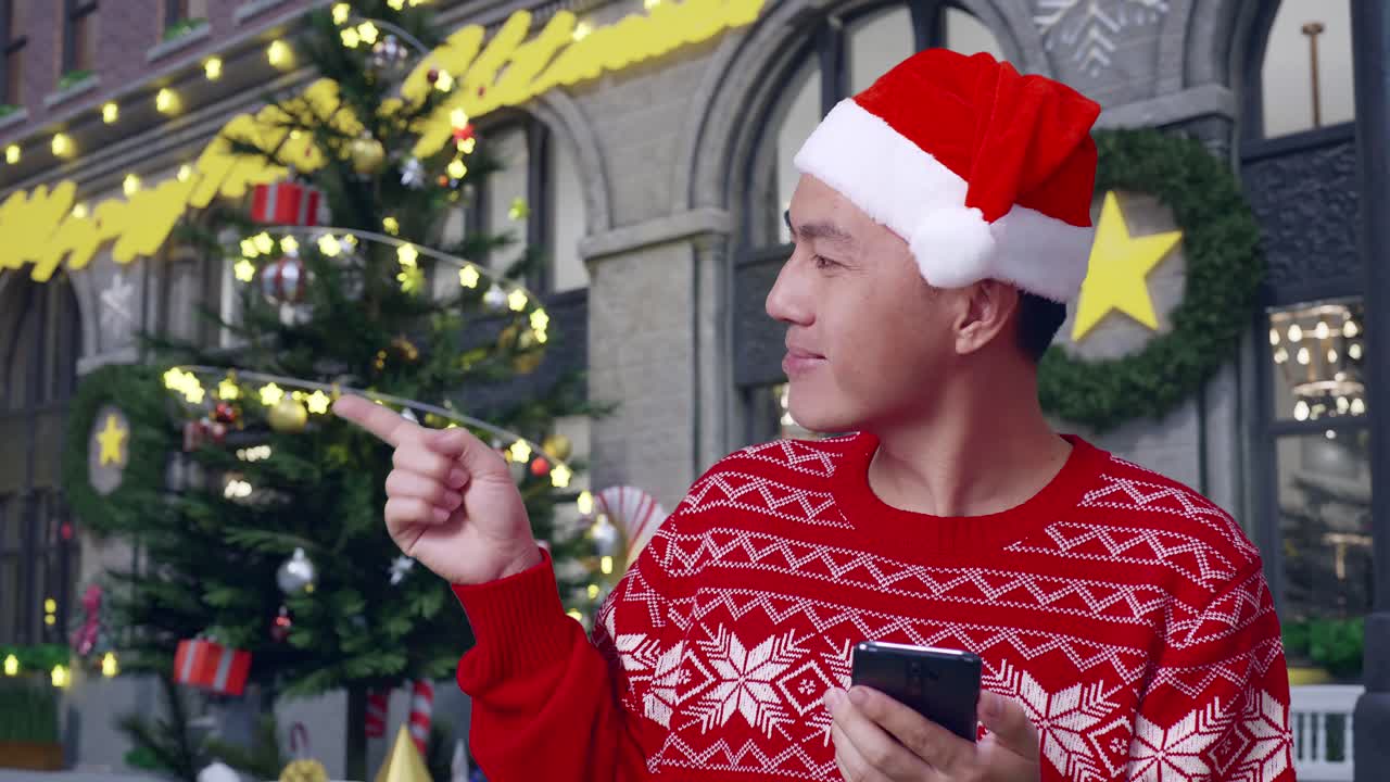 산타 의상을 입은 아시아인 남자가 스마트폰을 들고 크리스마스 밤에 서서 을 가리키며 미소 짓고 있습니다.