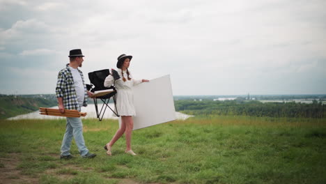 a man and woman stroll through a grassy field with a river in the background. the woman carries a canvas, and the man holds a drawing pad. both appear to be artists, with the man in a fedora shirt