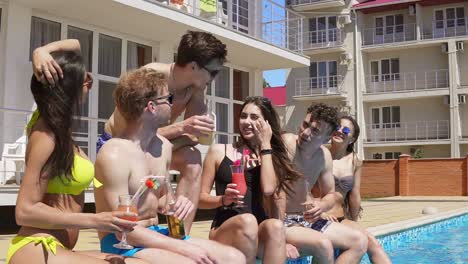 happy group of young friends hanging out with cocktails and chatting at the side of the pool in the summertime. pool party