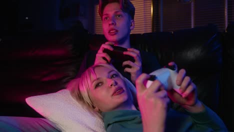 young caucasian couple playing video game with game pads while sitting and lying on sofa