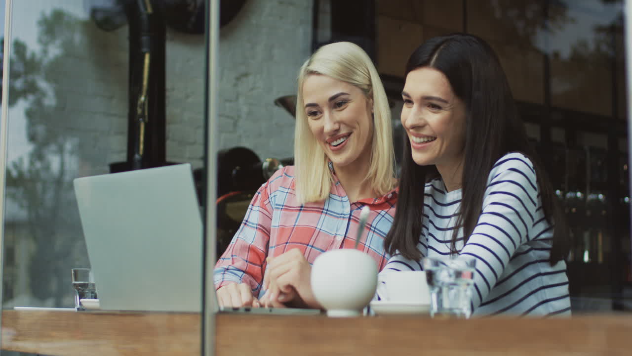 Deux jolies femmes assises devant un ordinateur portable, regardant quelque chose d'intéressant dans le café et en visioconférence