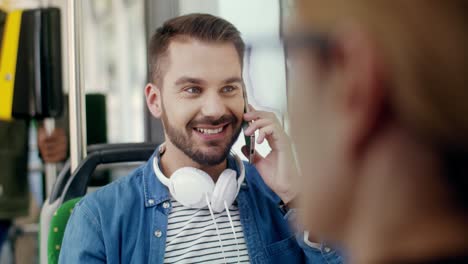 Close-Up-Of-The-Blurred-Rear-Businesswoman-And-Young-Attractive-Happy-Guy-With-Big-Headphones-On-His-Neck-Talking-Cheerfully-On-The-Mobile-Phone-On-The-Background