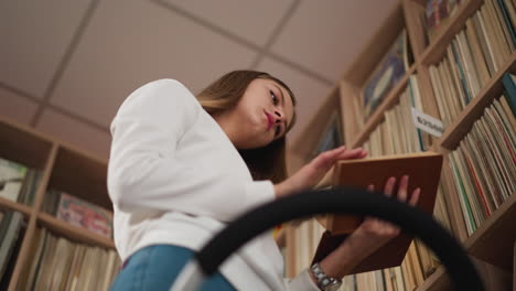 young woman chooses book from upper shelf. focused female student peruses book searching literature for scientific project. college library premise