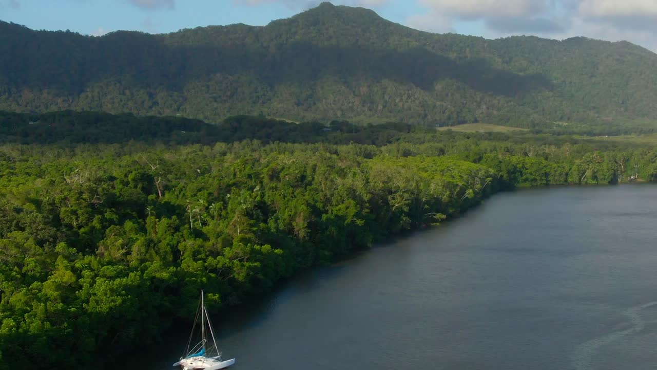 Two boats sit moored on the Daintree River next the the Daintree Rainforest. The camera pushes in and tilts up towards distant rainforest covered mountains.