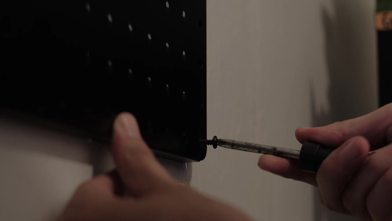 Close-up static shot of hands using a screwdriver to fasten a black pegboard to a wall during indoor installation work