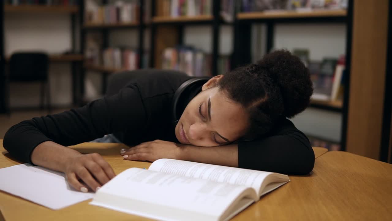 A female afro american student in black sweater, tired and fell asleep on a table with books and notes in the modern library. Education, people, student and learning concept. Front view