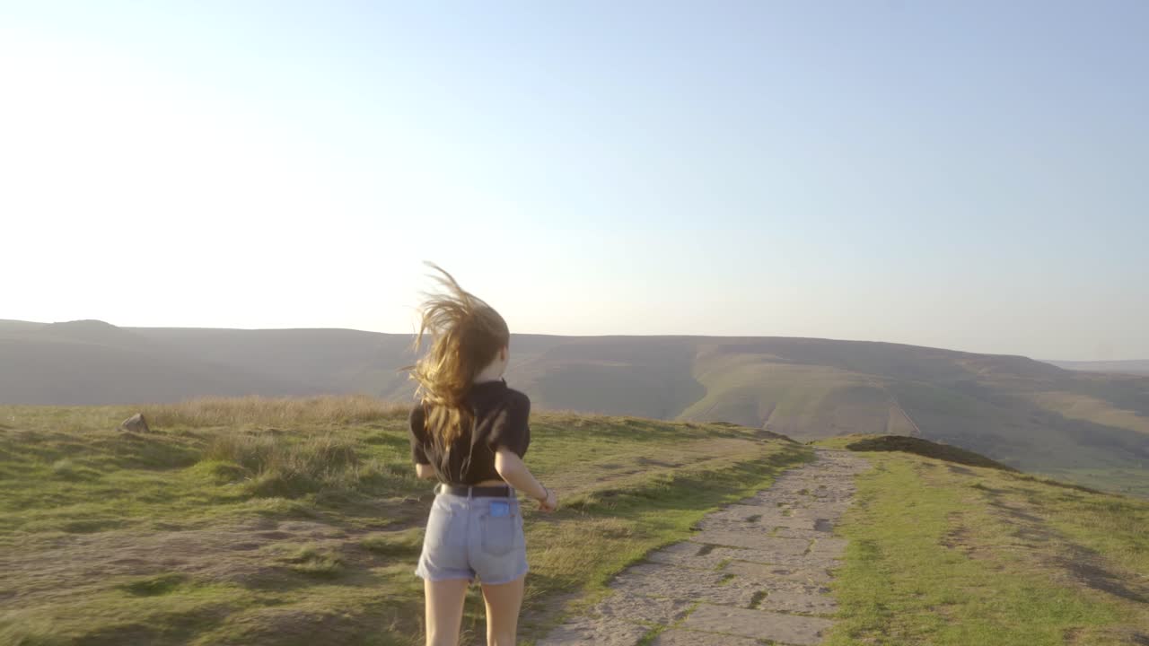 toma estabilizada de una joven rubia trotando a lo largo del camino en la parte superior de mam tor, castleton, distrito de los picos, inglaterra con vistas a verdes colinas y cielos azules