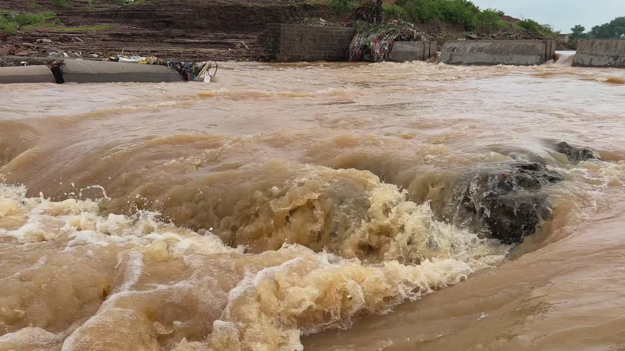 Static shof of intense flooding in a river and turbulent, muddy water flowing rapidly in higher volume and swift current, submerged concrete structures indicating a bridge or dam overwhelmed by flood