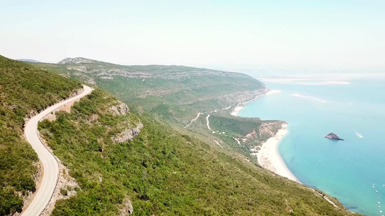 Breathtaking aerial view of a winding road traversing the lush green hills of Parque Natural da Arrábida, overlooking the stunning coastline and turquoise waters of the Atlantic Ocean in Portugal