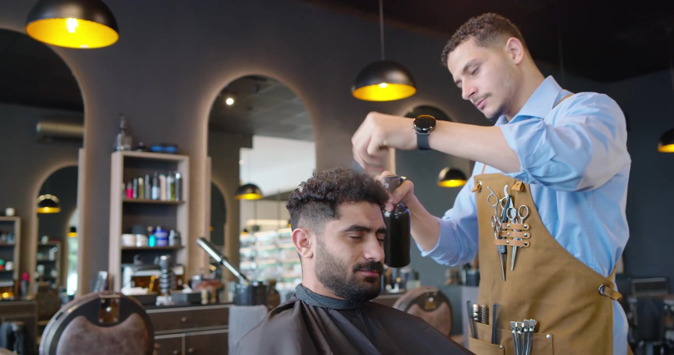 En afroamerikansk barber rækker ud efter en sort sprayflaske, sprayer og afslutter klipningen i salonen.
