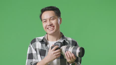 손에 카메라를 들고 미소 짓고 스튜디오에서 녹색 화면 배경에 서있는 아시아 사진작가의 클로즈업