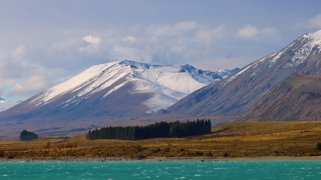 Wide panning shot reveals turquoise Lake Tekapo, snow-covered Southern Alps, autumn foliage, and pine forest under daylight with soft, natural lighting